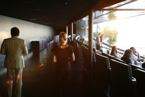 Menschen sitzen in zwei blauen Ledersessel-Reihen bei geöffneter Fensterfront und schauen Richtung Seebühne