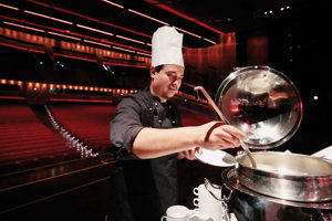 Ein Koch schöpft aus einem großen Topf, im Hintergrund die rote Reihenbestuhlung des Großen Saals im Festspielhaus Bregenz