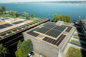 Luftaufnahme über das Werkstattbühnen-Dach mit großer Fotovoltaik-Anlage und dem Bodensee im Hintergrund