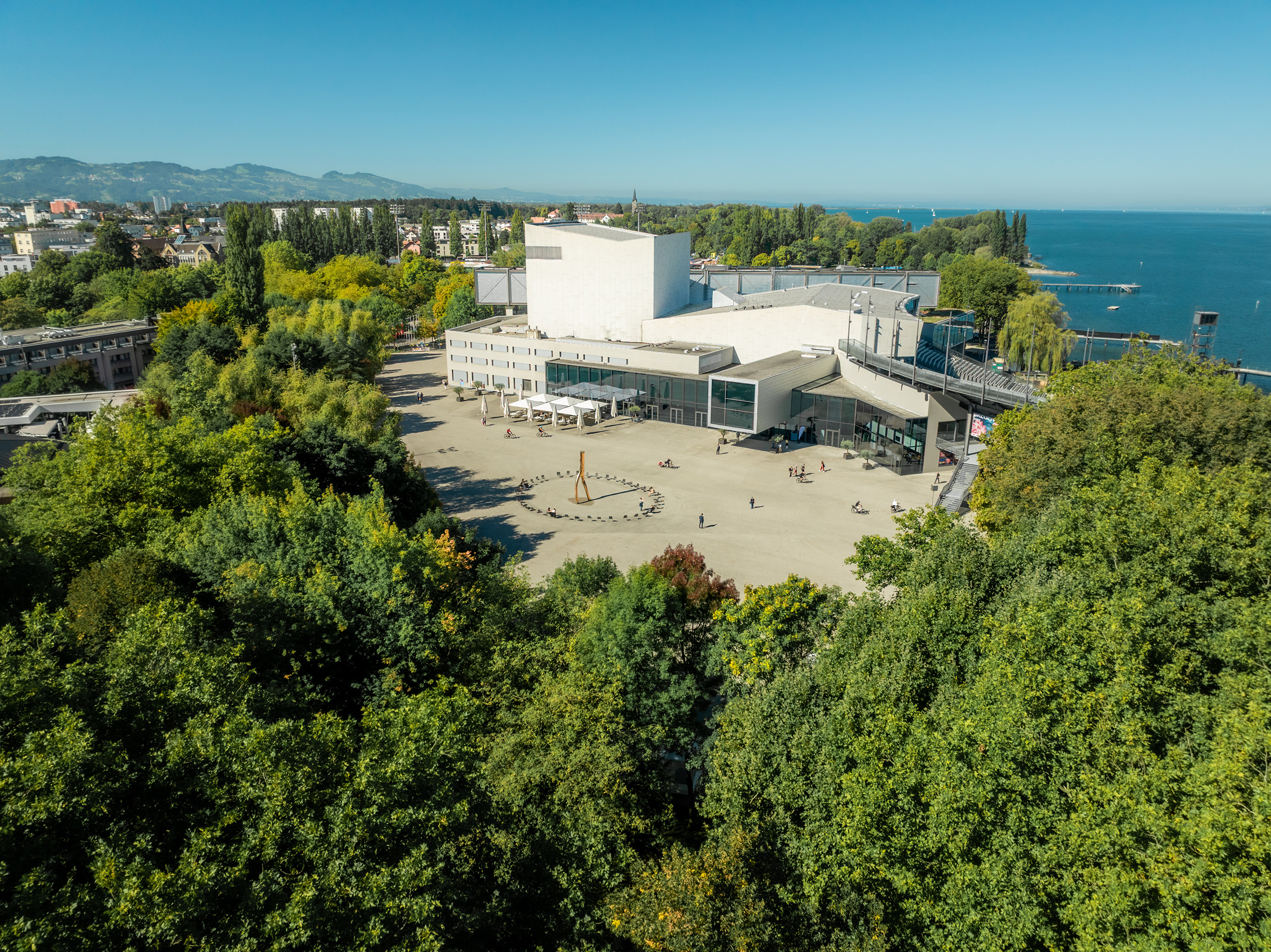 Drohnenaufnahme des Festspielhauses Bregenz mit vielen Bäumen und dem Bodensee im Hintergrund © bobdo