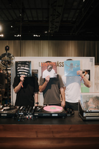 Drei Personen stehen hinter DJ-Pult und halten Schallplatten vor ihre Gesichter, im Hintergrund eine Eventfläche mit Lichttechnik.