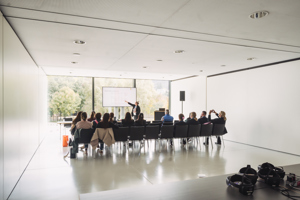 Kongressteilnehmer:innen sitzen in schwarzen Stuhlreihen vor einem Großbild-TV in einem weißen Raum