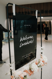 Schwarzes Schild mit Schrift "Welcome, Lovebirds!" an einem Gestell mit Blumen dekoriert.