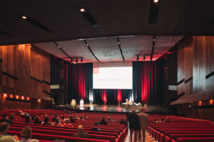 Kongressteilnehmer:innen sitzen in roten Stuhlreihen vor einer Bühne mit Podium im Großen Saal