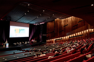 aufsteigendes Auditorium mit roten Stuhlreihen und schwarzen Tischen vor großer Leinwand