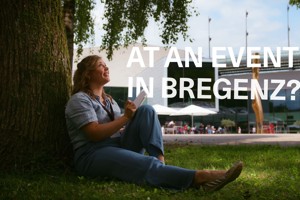 A woman is sitting, leaning against a tree, in front of the Festspielhaus Bregenz.