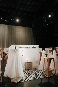 Mehrere Brautkleider hängen und stehen auf einer Ausstellungsfläche mit Teppich und Möbeln.